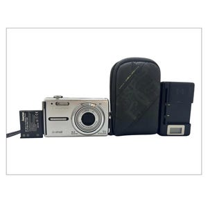 May include: A silver Olympus X-745 digital camera with a black lens, a black carrying case, a black battery charger, and a black battery. The camera has a 6.0 megapixel resolution and 6x optical zoom.