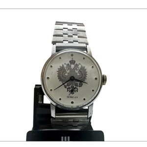 Puede incluir: Un reloj de pulsera plateado con una esfera redonda que presenta un emblema de águila bicéfala y la palabra cirílica "Победа". El reloj tiene una correa de metal con eslabones y una pequeña corona. El reloj se muestra en un soporte negro.