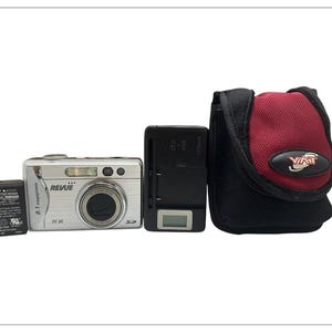 May include: A silver digital camera with a black lens, a battery, a charger, and a black and red carrying case. The camera is labelled "Revue" and "8.1 megapixels". The carrying case has the word "YAT" on it.