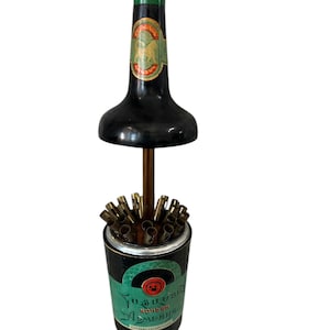 Könnte beinhalten: Ein Vintage-Zigarettenhalter in Schwarz und Grün in Form einer Flasche. Der Halter hat einen grünen Deckel und ein Etikett mit Text in einer Fremdsprache. Die Basis ist zylindrisch und enthält mehrere Zigarettenhalter.