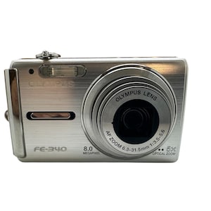 May include: Silver Olympus FE-340 digital camera. The camera has a lens with the text "Olympus Lens" and "AF Zoom 6.3-31.5mm 1:3.5-5.6". It also has "8.0 Megapixel" and "5x Optical Zoom" printed on the front.