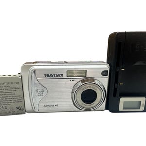 Pode incluir: Câmera digital Traveler Slimline X6 prateada com resolução de 6,2 megapixels. Inclui carregador preto e bateria de íon de lítio. A câmera possui lente com zoom 3x e uma pequena tela LCD.