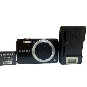 May include: A black Samsung digital camera with a silver lens ring, a battery, and a charger. The camera has a 5x zoom and the battery is labeled BP70A. The charger has an LCD screen.