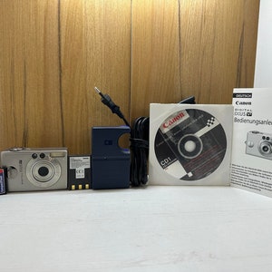 Cámara digital Canon Ixus V2 AiAf, objetivo zoom de 2 MP, compacta, batería de ion de litio, cargador de batería, tarjeta de memoria de 64 Mb, CD y libro