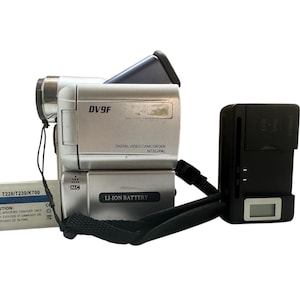 Peut inclure: Un caméscope numérique argenté avec une sangle noire et un chargeur noir avec un écran numérique. Le caméscope a un objectif noir et un corps argenté avec le texte "DV9F" et "LI-ION BATTERY". Le chargeur a le texte "For T226/T230/K700" et une étiquette d'avertissement.