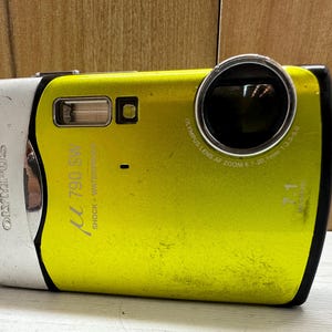 May include: A yellow Olympus &mu; [mu] 790 SW digital camera with a silver front panel and black lens. The camera has the text "Olympus" and "&mu; 790 SW" printed on the front. The text "Shock + Waterproof" is printed on the side of the camera. The lens has the text "Olympus Lens AF Zoom 6.7-20.1mm 1:3.5-5.0" printed around it.