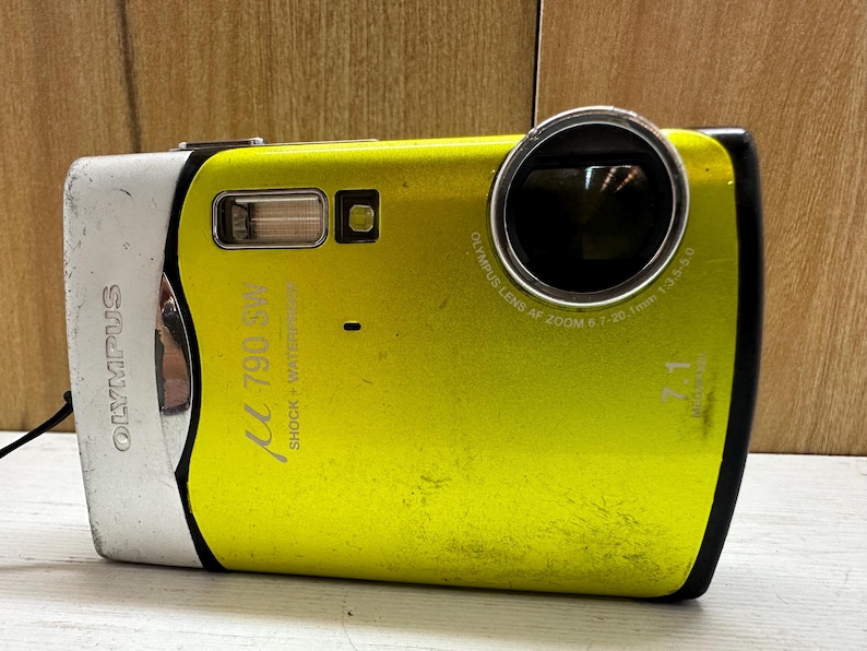 May include: A yellow Olympus &mu; [mu] 790 SW digital camera with a silver front panel and black lens. The camera has the text "Olympus" and "&mu; 790 SW" printed on the front. The text "Shock + Waterproof" is printed on the side of the camera. The lens has the text "Olympus Lens AF Zoom 6.7-20.1mm 1:3.5-5.0" printed around it.