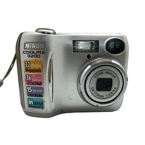 Peut inclure: Un appareil photo numérique Nikon Coolpix 3200 argenté avec une dragonne. La face avant présente un objectif Zoom Nikkor 5.8-17.4mm 1:2.8-4.9, un flash et une étiquette indiquant 3.2 mégapixels effectifs, zoom optique 3x, 15 modes scène et mémoire interne.