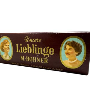 Puede incluir: Una caja marrón vintage con letras doradas y dos retratos ovalados de mujeres. El texto dice "Unsere Lieblinge M. Hohner" y "Made in Germany - Fabriqué en Allemagne".