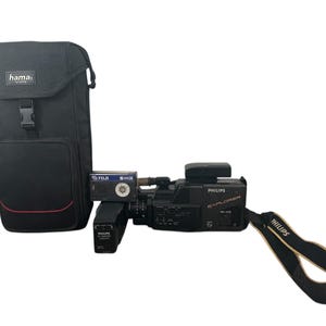 Puede incluir: Videocámara VHS Philips Explorer negra con una funda de transporte negra y una correa de muñeca negra y beige. La videocámara tiene una batería y una cinta insertada. La funda de transporte tiene el texto "hama".
