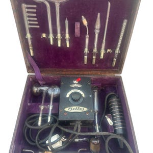 May include: A vintage Helios violet ray medical device set housed in a brown case with purple velvet lining. The kit includes a black control box with a dial labelled 110-220 Volt, various glass vacuum electrodes of different shapes, and a black handle.
