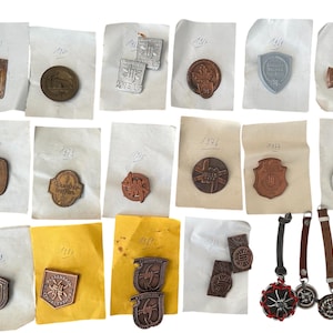 Puede incluir: Una colección de insignias y medallones metálicos antiguos, con diseños y textos en relieve, presentados sobre papel envejecido. Las insignias varían en forma, incluyendo escudos, círculos y rectángulos. Algunos están unidos a correas de cuero, en tonos bronce a plata.