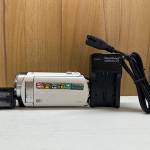 Pode incluir: Uma câmara de vídeo branca com uma bateria preta e um carregador preto com o texto "Blumax Charger Kit".