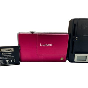 Op de afbeelding: Een magenta Lumix digitale camera met een zwarte batterij en oplader. De batterij is voorzien van een Panasonic label en de oplader heeft een LCD-scherm. De camera en oplader staan op een wit oppervlak.