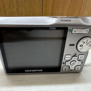 May include: A silver Olympus digital camera with a black screen and buttons. The camera has a zoom lens and a built-in flash. The camera is in good condition and appears to be used.