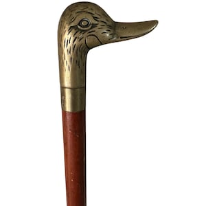 Pode incluir: Uma bengala com um cabo de latão polido em forma de cabeça de pato com detalhes de penas gravados. O cabo está fixado a um eixo de madeira cilíndrico e liso com um acabamento castanho quente, ligado por uma simples banda de latão.
