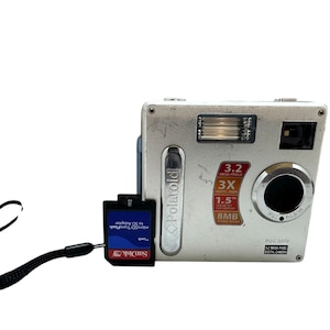May include: Silver Polaroid digital camera with a black strap and a black and red SanDisk SD card adapter. The camera has a 3.2-megapixel resolution, 3x zoom, and a 1.5-inch screen.