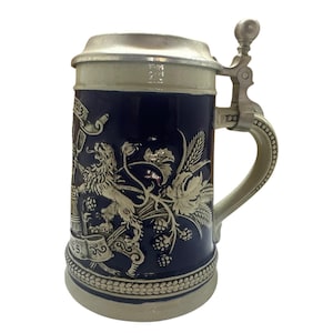May include: A ceramic beer stein with a dark blue body and a decorative relief design. The design includes a lion, wheat, and grapes. The stein has a hinged metal lid and handle. The base and lid are a light beige color.