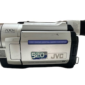 Puede incluir: Una videocámara MiniDV JVC plateada y negra con lente de zoom digital 700x, etiqueta de high-band digital video camera y placa de 520 lines super high-band processor. El dispositivo tiene un cuerpo rectangular con un visor lateral.