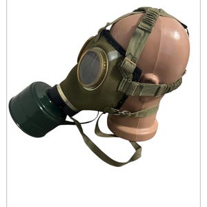 Könnte beinhalten: Eine olivgrüne Gasmaske mit einer kreisförmigen Linse und einem dunkelgrünen Filter. Die Maske wird mit verstellbaren Riemen befestigt und auf einem Schaufensterkopf präsentiert. Die Maske hat eine Vintage- oder Militärästhetik.