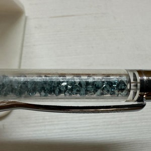 Puede incluir: Un bolígrafo transparente con un capuchón plateado y un clip plateado. El bolígrafo está lleno de pequeños cristales azul oscuro.