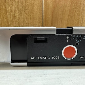 Può includere: Una macchina fotografica Agfamatic 4008 nera e argentata con un pulsante del sensore rosso. La macchina fotografica ha una scala di distanza in piedi e metri.