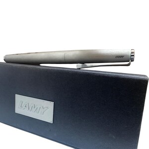 May include: A brushed silver Lamy fountain pen with a metallic clip resting on a rectangular black presentation box. The box features a silver-toned metal plaque with the embossed Lamy logo, and the pen barrel also displays the Lamy brand name.
