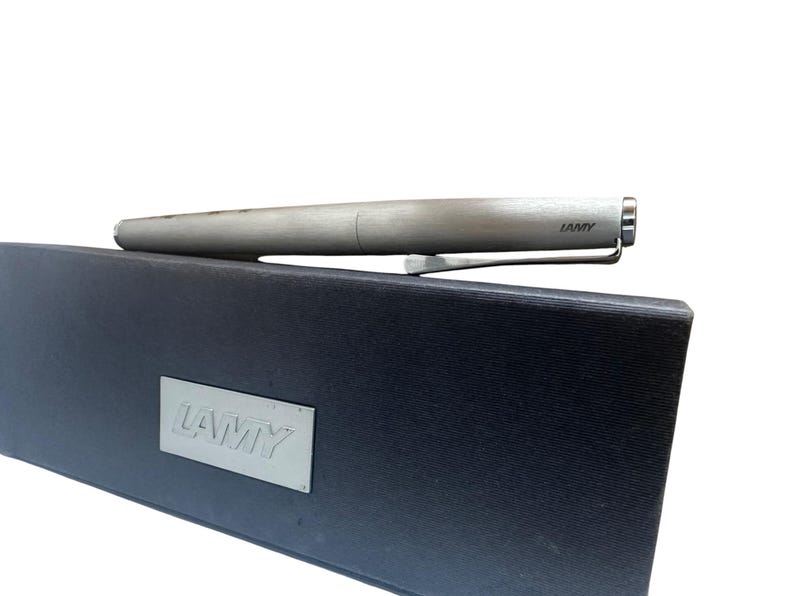 May include: A brushed silver Lamy fountain pen with a metallic clip resting on a rectangular black presentation box. The box features a silver-toned metal plaque with the embossed Lamy logo, and the pen barrel also displays the Lamy brand name.