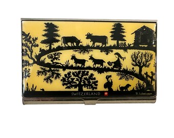 May include: A rectangular metal business card holder with a yellow background and a black silhouette design depicting a Swiss countryside scene. The design includes cows, goats, trees, and a small house. The word "SWITZERLAND" is printed on the holder.