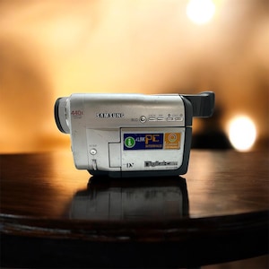 Peut inclure: Caméscope Samsung Digitalcam argenté avec des détails noirs. Il présente un zoom 440x, BLC et une interface PC. Le caméscope est posé sur une surface en bois sombre.