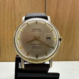 May include: A gold-toned wristwatch with a black leather band. The watch face is white with black markings and the text "ORIMA EXTRA PLATE" and "SUPER AUTOMATIQUE SHOCKPROOF ANTIMAGNETIC".