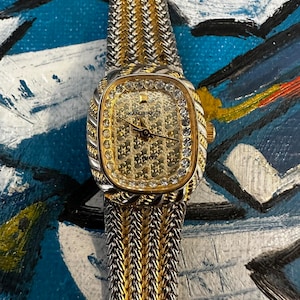 May include: A gold and silver watch with a square face and a diamond-encrusted bezel. The watch face is gold with a white dial and black hands. The watch band is a two-tone gold and silver chain link style.