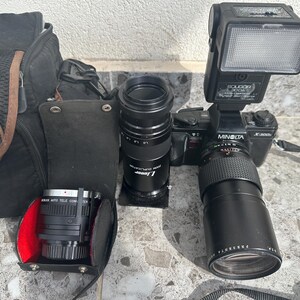 以下が含まれることがあります： 黒いミノルタX-3000カメラ、黒いフラッシュ、黒いレンズ、黒い望遠レンズ。カメラは黒いカメラバッグのあるテーブルの上に置かれています。