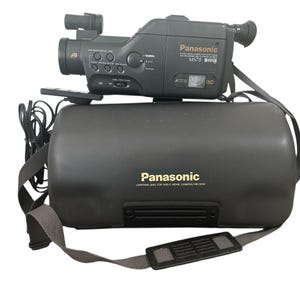 Op de afbeelding: Een vintage Panasonic MS70 camcorder in donkergrijs, rustend op een bijpassende draagtas. De camcorder heeft meerdere knoppen en een zoeker. De tas heeft een schouderriem en het Panasonic-logo.