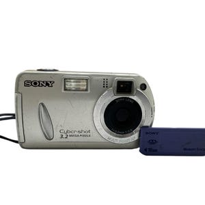 Könnte beinhalten: Silberne Sony Cyber-shot Digitalkamera mit 3,2 Megapixeln, schwarzem Objektiv und schwarzer Handschlaufe. Neben der Kamera befindet sich ein blauer 16-MB-Memory Stick. Das Sony-Logo ist sichtbar.