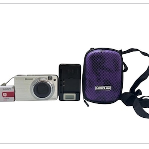 May include: A silver digital camera, battery, charger, and a purple camera bag with a black strap. The camera bag has the text "Camera-bag" on the front. The battery is red and white with the brand name "SONY".