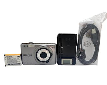 May include: A silver Fujifilm digital camera with a black lens, a battery, a charger, and a USB cable. The camera has the text "FUJIFILM" and "14 MEGA PIXELS" printed on it. The battery is yellow and white.