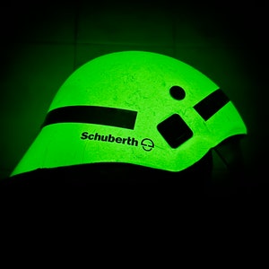 Op de afbeelding: Een lichtgevende groene helm met het woord "Schuberth" aan de zijkant. De helm heeft een zwarte streep en een paar zwarte vierkanten. De afbeelding staat tegen een donkere achtergrond.