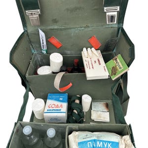Puede incluir: Un botiquín de primeros auxilios de lona verde con una variedad de suministros médicos, incluidos vendajes, gasa, bolas de algodón y botellas de líquido. El botiquín está abierto y el contenido es visible. El botiquín tiene una etiqueta que dice "Acetylsal" y otra etiqueta que dice "Soda Bikarbonat".