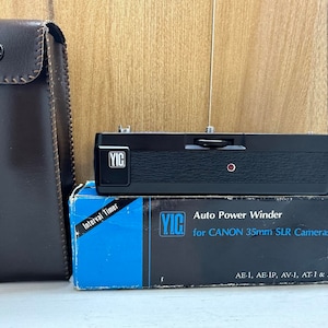 Puede incluir: Un enrollador automático de energía negro y plateado para cámaras réflex Canon de 35 mm. El enrollador está en una caja con el texto "YIG Auto Power Winder for CANON 35mm SLR Cameras AE-1, AE-IP, AV-1, AT-1 & A-1" impreso en ella.