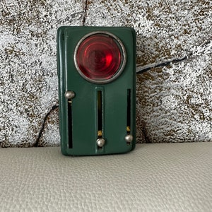 Może przedstawiać: Zielona metalowa zabawka z czerwoną szklaną soczewką i trzema srebrnymi gałkami. Zabawka to sygnalizacja awaryjna w stylu vintage.