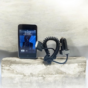 Puede incluir: Un iPod con una pantalla negra que muestra un gráfico azul y la hora 3:02. Un cargador de coche negro y un cable de carga negro en espiral están junto al iPod. Los artículos están sobre una superficie gris claro.