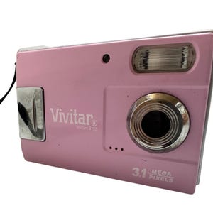 May include: A pink Vivitar digital camera with a silver lens and flash. The camera has a wrist strap and the text "Vivitar" and "3.1 Mega Pixels" are visible on the front.
