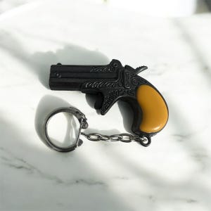 Op de afbeelding: Zwarte miniatuur pistool sleutelhanger met een gele handgreep en zilveren ketting. Het pistool heeft gedetailleerde gravures. De sleutelhanger is bevestigd aan een zilveren ring en ligt op een witte marmeren ondergrond.