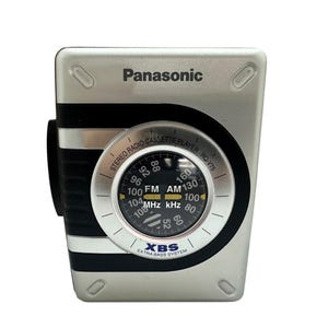 Puede incluir: Reproductor de casetes de radio estéreo Panasonic plateado. El dispositivo presenta un dial circular con indicadores de frecuencia FM y AM. El texto "XBS EXTRA BASS SYSTEM" es visible. El número de modelo es RQ-V75.