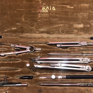 May include: A vintage set of drafting compasses in a brown leather case. The compasses are made of silver metal and have a black handle. The case is lined with brown velvet. The case has the word "Knia" and "Precision" written on it.
