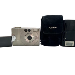 Może przedstawiać: Srebrny aparat cyfrowy Canon Digital IXUS 400 z czarną ładowarką do baterii, czarnym etui Canon i czarną lampą błyskową. Aparat ma obiektyw 2,8 mm. Logo Canon jest widoczne na etui.