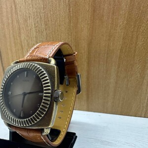 May include: A square-shaped, gold-toned wristwatch with a brown leather strap. The watch face has a dark brown background with silver hands and markers. The word "GUESS" is printed in silver on the watch face.
