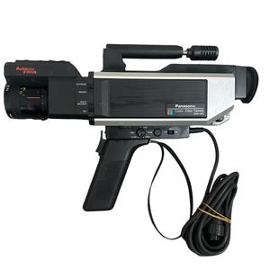 Könnte beinhalten: Eine Vintage Panasonic WV-50E Farbvideokamera. Die Kamera ist schwarz und silberfarben mit einem Griff, Objektiv und verschiedenen Bedienelementen. Sie hat ein schwarzes Kabel und einen Handgriff. Auf dem Objektiv steht "Auto Focus".