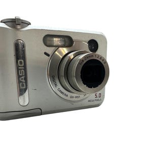 Peut inclure: Appareil photo numérique Casio argenté avec objectif et flash. Le mot "CASIO" est imprimé sur le côté et "DIGITAL CAMERA QV-R51" sur l'objectif. Il a une résolution de 5,0 mégapixels.